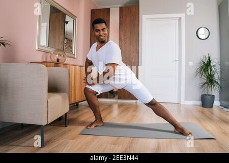 Vista frontale di un forte uomo afro-americano sorridente che fa affondo laterale esercitarsi a casa Foto Stock