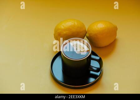 tè. Tè con una fetta di limone in una tazza nera su un piatto. Due limoni su sfondo giallo Foto Stock