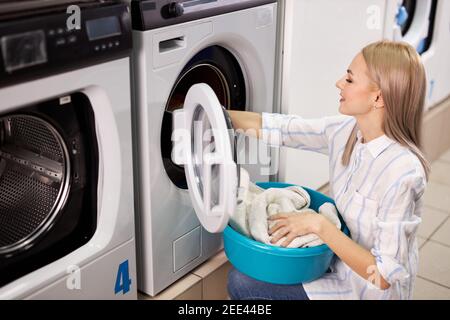 donna in casa di lavaggio smistamento vestiti puliti, facendo compiti domestici, donna tira fuori i vestiti dalla lavatrice, tenendo bacino. vista laterale Foto Stock