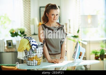 felice casalinga trendy di 40 anni in blusa di seta e pantaloni beige con ferro da stiro a vapore, cesto di vestiti lavati e asse da stiro nella casa moderna al sole Foto Stock