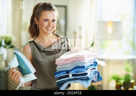 Ritratto di sorridente casalinga moderna di 40 anni in seta blusa e pantaloni beige con ferro da stiro a vapore e pile di vestiti stirati piegati in casa moderna in s. Foto Stock