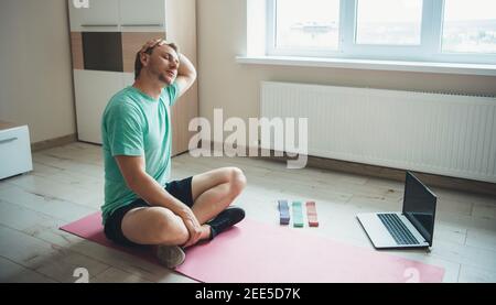 Stretching uomo caucasico è seduto sul pavimento in abbigliamento sportivo e utilizzando un computer portatile durante la sua sessione di fitness Foto Stock