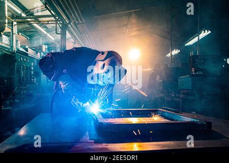 I due handymen che eseguono la saldatura e la molatura sul luogo di lavoro in officina, mentre le scintille "volano" tutto intorno a loro, indossano un lui protettivo Foto Stock