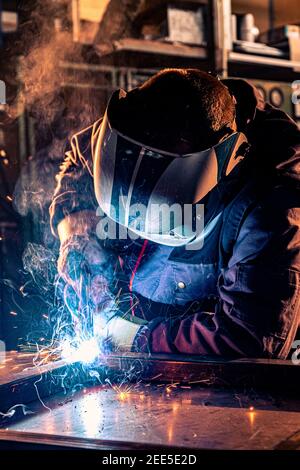 Primo piano di una saldatrice in officina. È vestito con una divisa da lavoro e ha un casco protettivo sulla testa mentre le scintille volano in tutte le direzioni. Foto Stock