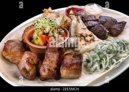 Carne alla griglia assortita, kebab, maiale, manzo, pollo. Con pane pita, cipolle e verdure alla griglia. Su una piastra bianca. Su sfondo nero. Foto Stock