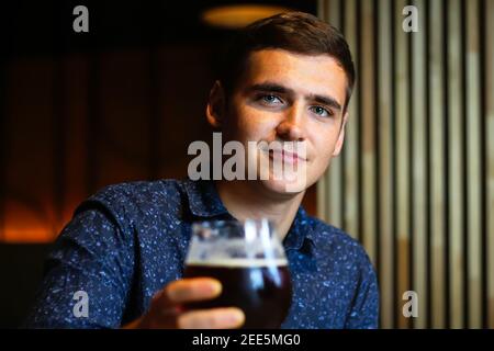 Bel giovane caucasico dai capelli castani che tiene un bicchiere di birra al ristorante. Concetto di persone, bevande, alcol e tempo libero - buon giovane uomo che beve Foto Stock