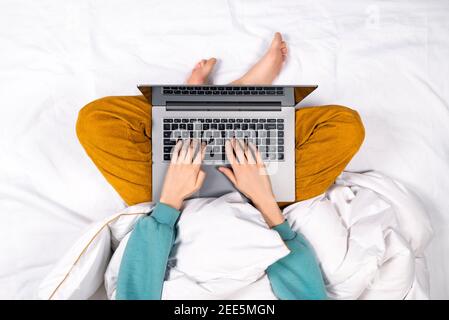 Vista dall'alto sulle mani femminili che lavorano su un computer portatile moderno dal letto a casa. Concetto di home Office o formazione online. Il laptop si trova sul grembo di una donna. Foto Stock