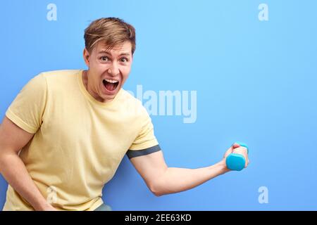 maschio non atletico che tiene in mano un piccolo dumbell, è difficile per lui sollevarlo, urlare, stare in piedi con la bocca aperta, allenarsi, pompare il mus del braccio Foto Stock