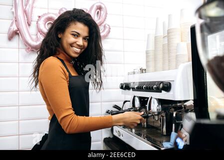 Vista laterale una sorridente e amichevole barista afroamericana fa caffè in tazza di carta per i visitatori del caffè utilizzando la macchina del caffè, in piedi dietro il bancone del bar e sorridente Foto Stock