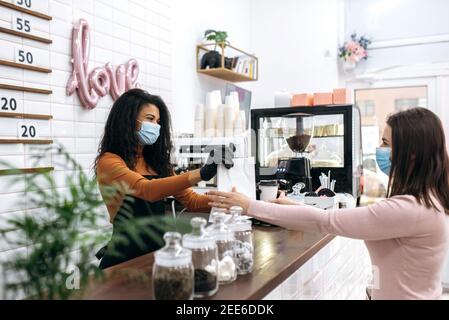 Barista afroamericano ragazza in uniforme, maschera medica e guanti protettivi dà una donna visitatore caffè in un bicchiere di cartone monouso e dessert in una borsa di carta per andare. Concetto di caffè in movimento Foto Stock