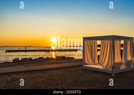 Patio con lettini e tende sulla spiaggia e come sagoma di persona sul molo in marina al tramonto sul mare Adriatico con Sunstar definito, Zaton, Croazia Foto Stock