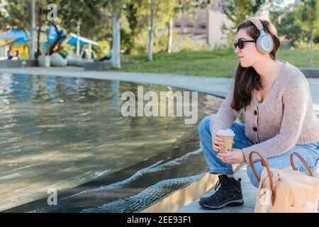 Giovane caucasica che ascolta musica con le cuffie mentre beve caffè in un parco Foto Stock