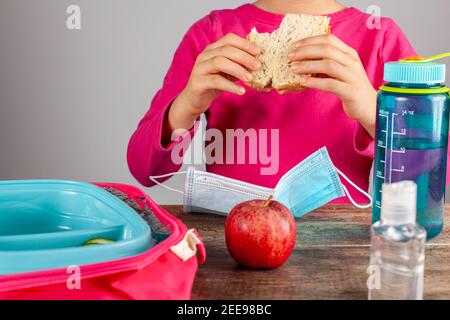 Immagine closeup con pranzo a scuola durante la riapertura graduale dopo la chiusura pandemica del COVID-19. Ragazza con maschera viso rimosso mangia sandw Foto Stock