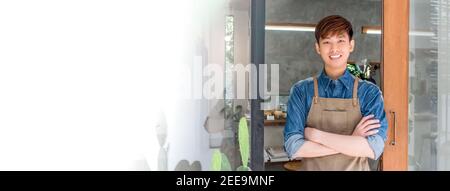 Giovane imprenditore asiatico uomo in piedi e incrociando le braccia alla porta anteriore del suo proprio caffè, banner panoramico con spazio di copia Foto Stock