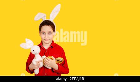 Ragazzo piccolo con orecchie conigliate che tengono le uova di Pasqua sul giallo sfondo Foto Stock