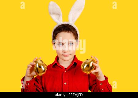 Ragazzo carino con orecchie conigliate che tengono le uova di Pasqua sul giallo sfondo Foto Stock