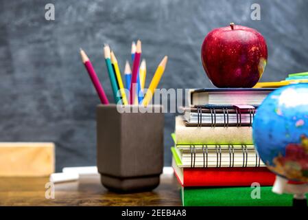 Stack di libri colorati, cancelleria e materiale didattico su scrivania in legno in classe con lavagna in background Foto Stock