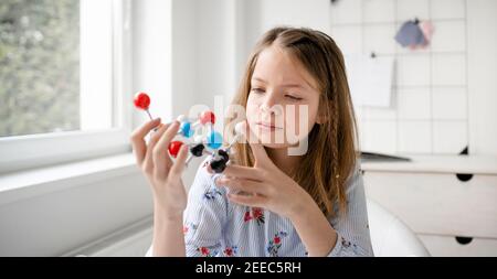 la scolastica abbastanza bionda sta imparando la chimica con l'aiuto di un modello di molecola ed è seduto al tavolo Foto Stock