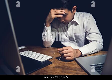 Stressato giovane uomo stanco che sente mal di testa mentre lavora straordinario su il suo computer portatile di notte finendo il suo progetto Foto Stock