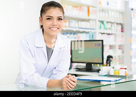 Sorridente bella giovane donna asiatica farmacista in camice bianco al banco in farmacia (farmacia o farmacia) Foto Stock