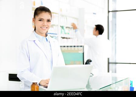 Giovane donna asiatica sorridente farmacista in abito bianco uniforme lavoro su computer portatile in farmacia o negozio di farmacia Foto Stock