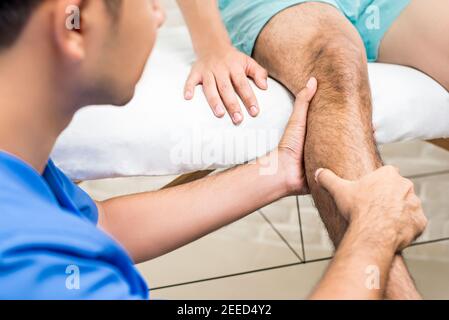 Medico (terapista) che tratta la gamba del paziente maschile mentre si siede sul letto in clinica - concetto di terapia fisica Foto Stock