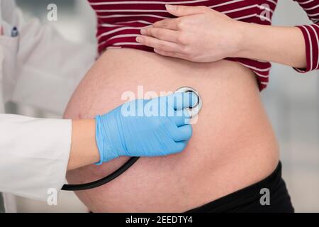 Pancia di giovane donna incinta controllata da un medico utilizzando uno stetoscopio. Messa a fuoco selettiva e primo piano. Concetto di gravidanza e assistenza sanitaria. Foto Stock