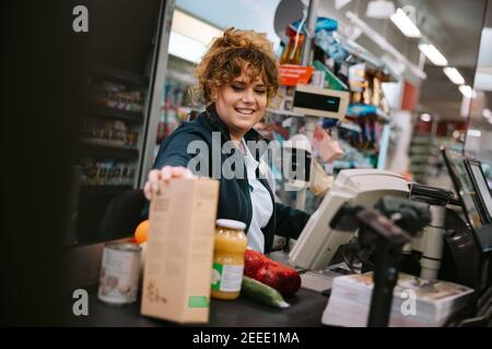 Accogliente cassiere che passa i prodotti attraverso il lettore di codici a barre al supermercato. Donna che lavora al registratore di cassa del negozio di alimentari. Foto Stock