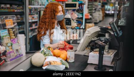 Impiegato donna con maschera in un supermercato che lavora al banco cassa. Donna cassiere che scannerizzare gli articoli della drogheria per il cliente durante la pandemia. Foto Stock