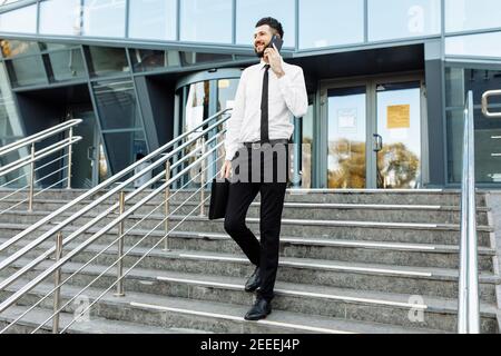 Uomo d'affari di stile in un vestito cammina lungo i gradini e. parla al telefono sulla strada vicino all'ufficio edificio Foto Stock