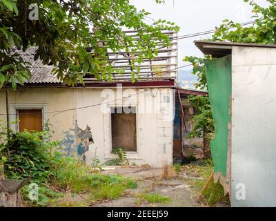 Una vecchia casa abbandonata ad un piano con tetto distrutto e rotto finestre ed erba che crescono attraverso il pavimento di calcestruzzo nel iarda Foto Stock