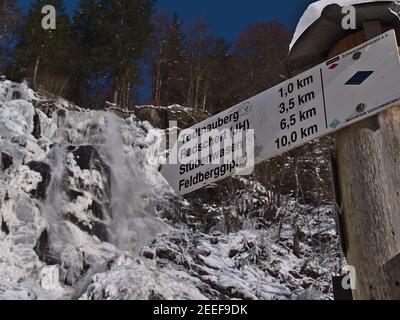 Segnaletica con destinazioni e distanze di trekking di fronte alla famosa cascata Todtnauer Wasserfälle in inverno con ghiaccio e neve. Concentrarsi sul centro. Foto Stock