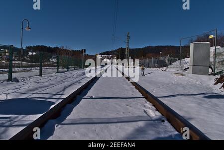 Ridotta prospettiva di binari ferroviari innevati con cavetto sopraelevato circondato da recinzioni nella soleggiata giornata invernale con cielo blu. Foto Stock