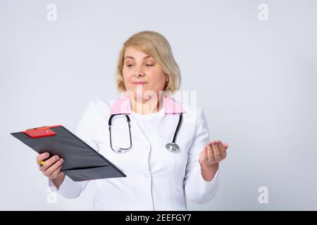 Medico anziano bello, donna con capelli biondi, indossando uniforme medica, guardando su cartella foglio con prescrizione. Espressione confusa con le braccia e. Foto Stock