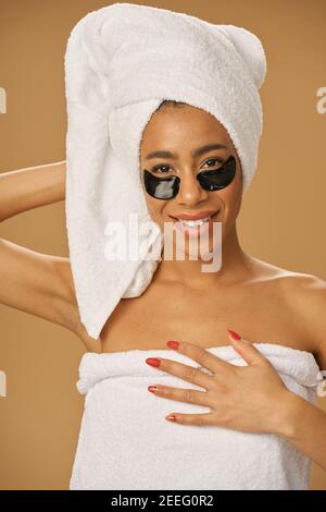 Giovane donna allegra con asciugamano che sorride alla macchina fotografica, posando con applicate macchie di bellezza nere sotto gli occhi isolati su sfondo beige. Bagno routine. Concetto di trattamento sotto gli occhi Foto Stock