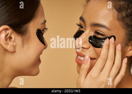 Primo piano ritratto di belle giovani donne che si guardano l'un l'altro, applicando nero idrogel sotto gli occhi macchie sulla pelle del viso, in posa isolato su sfondo beige. Routine di cura della pelle, concetto di diversità Foto Stock