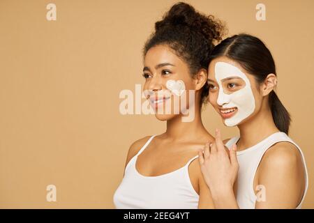 Ritratto di due giovani donne, amiche sorridenti mentre si posano insieme con maschere facciali su sfondo beige isolato. Cura della pelle, concetto di bellezza Foto Stock