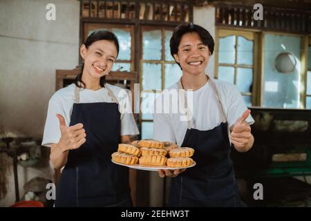 giovane coppia di produttori di torte che indossano grembiuli con pollici in su come un piatto di biscotti cotti al forno in cucina come uno sfondo Foto Stock