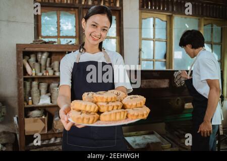 sorridente ragazza che fa una torta che indossa un grembiule che porta uno piatto di torta con un uomo che apre il forno come uno sfondo Foto Stock