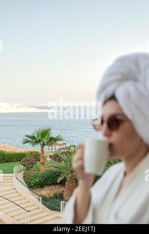 Immagine sfocata di donna con bicchieri in un accappatoio e. un asciugamano beve caffè Foto Stock