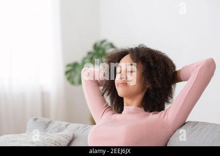 Calma serena giovane casalinga afroamericana che si rilassa sul divano di comfort a casa con gli occhi chiusi Foto Stock