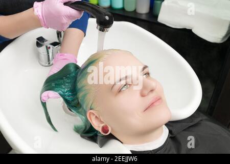 Hairstylist mani in guanto protettivo lavando i capelli verdi di bella giovane donna con shampoo in doccia in speciale salone di bellezza lavello. Lavoro capelli Foto Stock