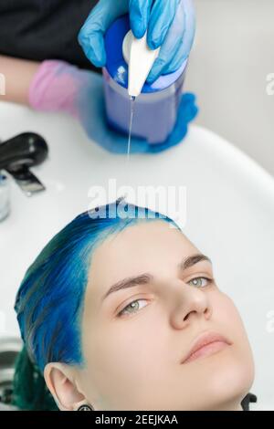 Vista ravvicinata delle mani del parrucchiere nei guanti strizza lo shampoo e. balsamo dal tubo alla testa della donna con i capelli blu lavando i capelli nel lavandino Foto Stock