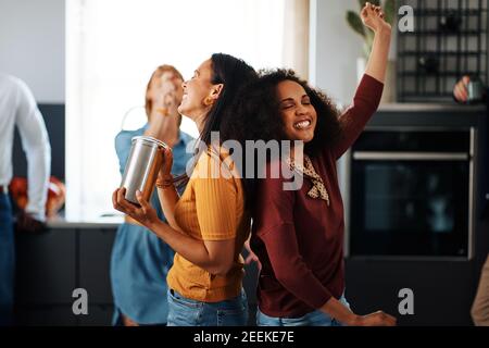 Gruppo ridente di diversi giovani amici che ballano e cantano durante una festa in cucina di un amico Foto Stock