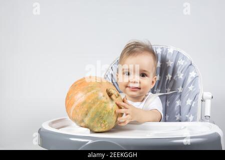 Un bambino piccolo carino che tiene una zucca fresca cruda su una sedia alta su uno sfondo bianco. Vitamine per i bambini negli alimenti. Foto Stock