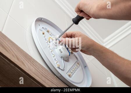 Un uomo sta usando un cacciavite per montare una lampada LED nel bagno alla parete. Riparazione e miglioramento dell'alloggiamento. Foto Stock