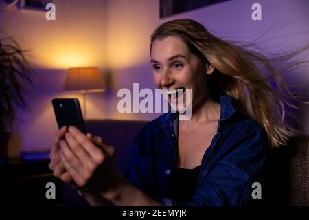 scioccato sorpresa giovane donna seduta sul divano e utilizzando il telefono cellulare. soffiare i capelli Foto Stock
