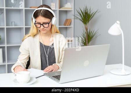 La ragazza studentessa in cuffia ascolta le lezioni dell'insegnante online utilizzando un computer portatile. Una giovane donna in occhiali seduto a casa scrive una lezione in una nota Foto Stock