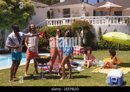 Un gruppo variegato di amici che fanno barbecue e parlano a una festa in piscina, che si può appendere, bere birra, fare un brindisi e rilassarsi all'aperto in estate. Foto Stock