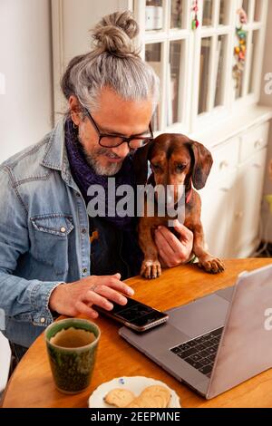 Bell'uomo che lavora a casa con il computer portatile sul tavolo insieme al suo cane carino; concetto: Stretto rapporto tra cane e uomo Foto Stock